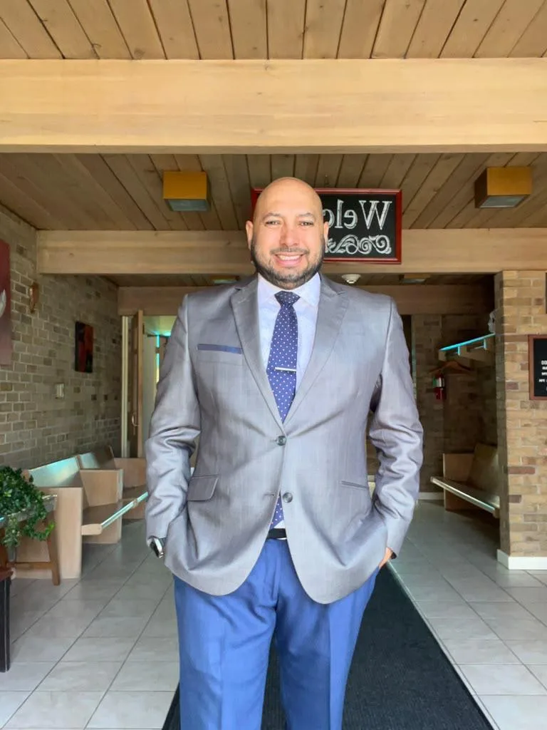José, owner and barber at Jose's Barber Shop in Portage Park, Chicago, smiling in a gray suit jacket and navy tie