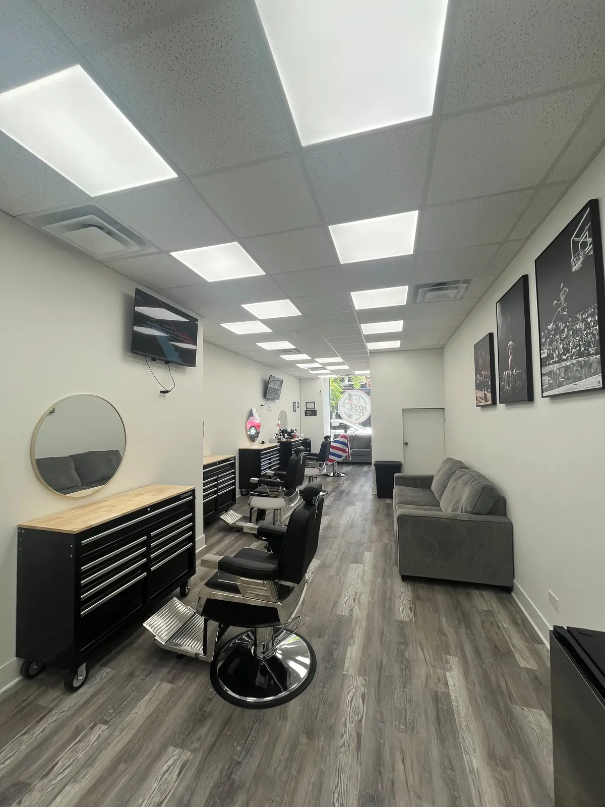 The floor of Jose's Barber Shop looking toward Milwaukee Ave — black barber chairs with chrome bases beside wood-topped tool chests, a round wall mirror, a gray waiting sofa under framed black-and-white sports photographs, and the shop's logo on the front window