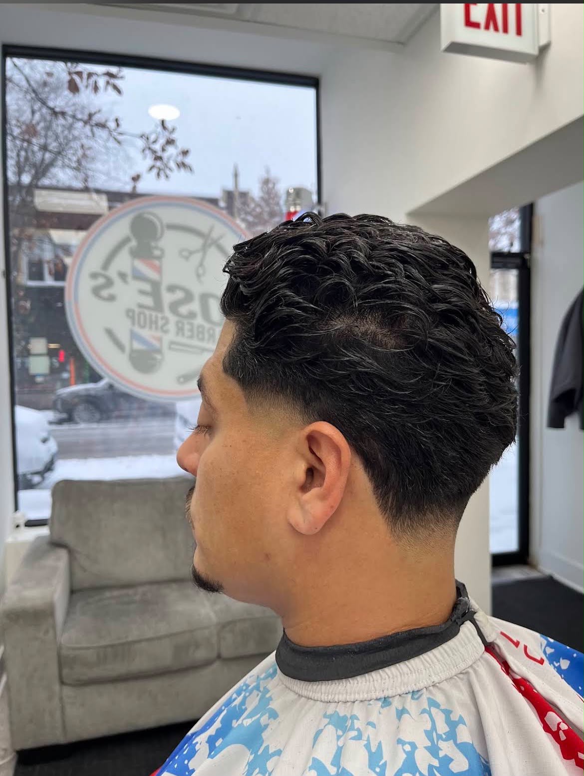 Cabello oscuro y rizado, largo arriba y degradado bajo en los lados y la nuca, fotografiado de perfil frente a la ventana y el logo de la barbería