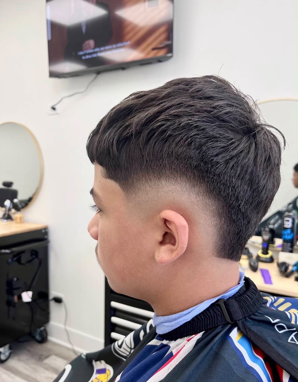 Un corte con textura, flequillo recto y un fade medio que baja detrás de la oreja, dejado más largo en la nuca, con la televisión de pared y un espejo redondo al fondo