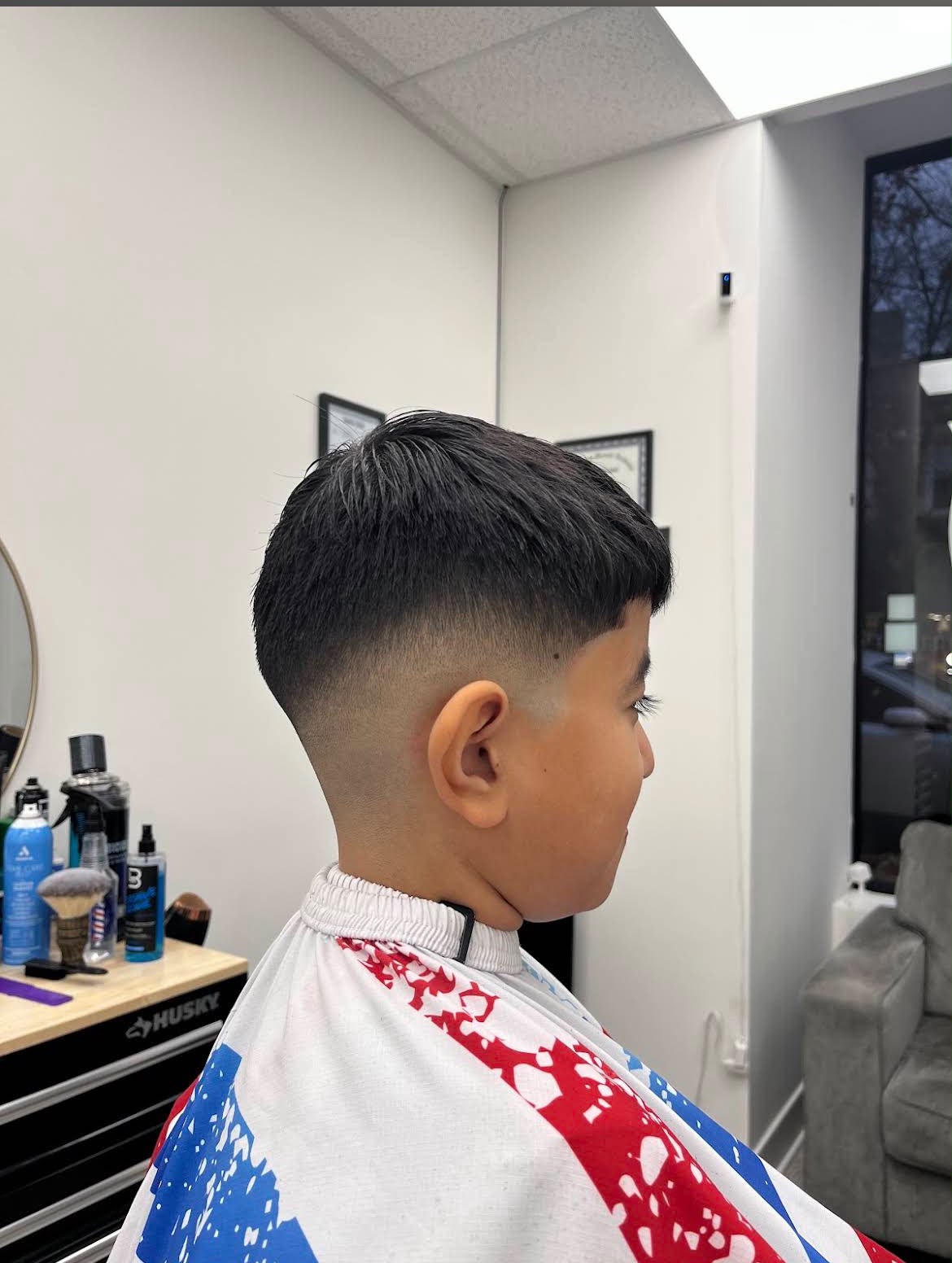 El corte terminado de un niño — cabello oscuro con textura, abundante arriba y con flequillo suave, difuminado en un taper fade limpio alrededor de la oreja, bajo la capa roja, blanca y azul de la barbería