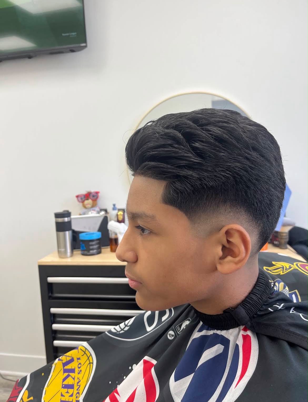 El cabello oscuro y abundante de un cliente joven, peinado hacia atrás arriba, con un delineado nítido en la sien y un taper bajo en los lados, frente al gabinete de herramientas y los productos