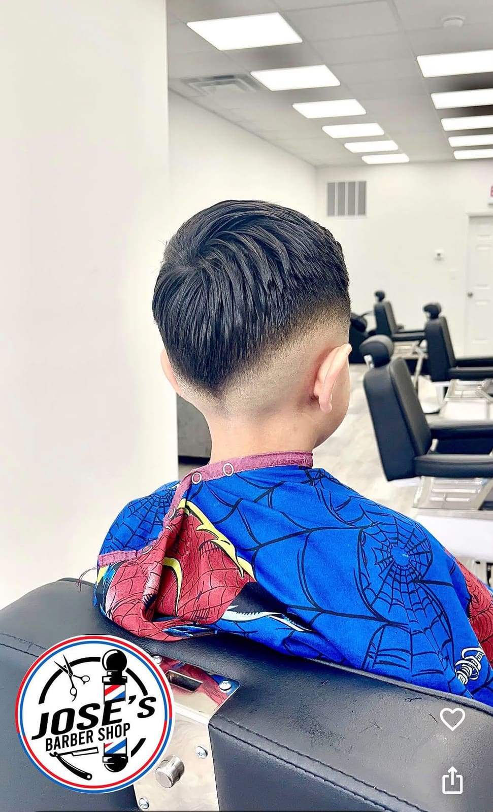 La parte de atrás del corte recién hecho de un niño — cabello con textura arriba y degradado hasta la piel en los lados y la nuca, con una capa de Spider-Man y la fila de sillas de barbero al fondo