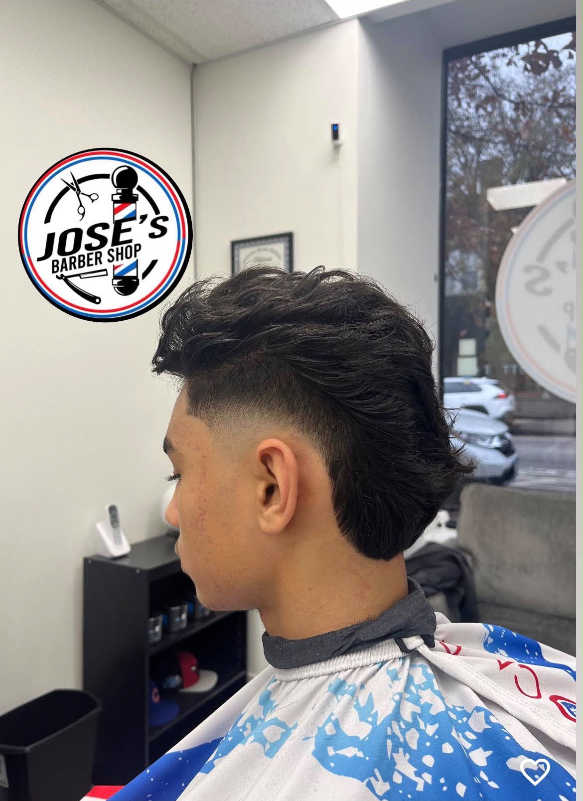 Cabello ondulado y largo, abundante arriba y en la parte de atrás, trabajado contra un skin fade alto y un delineado marcado en la sien, fotografiado junto a la ventana de Jose's Barber Shop