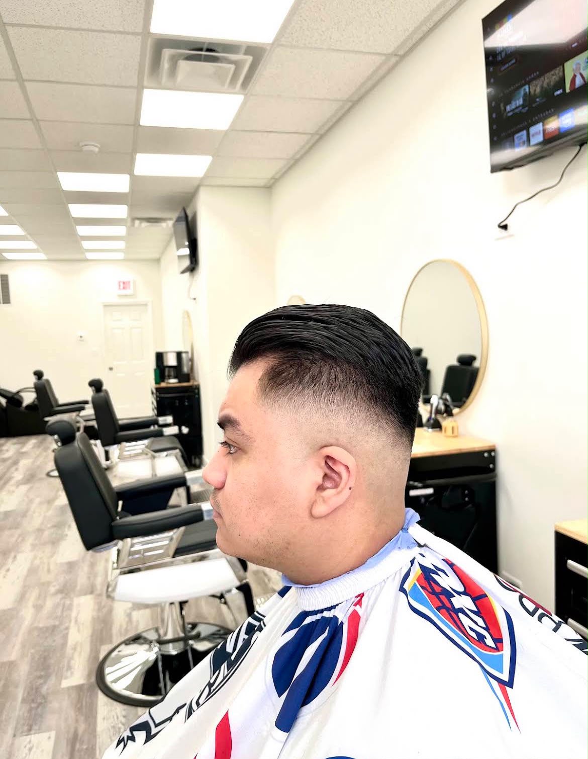 El cabello de un cliente peinado hacia atrás sobre un skin fade alto, de perfil en el salón de Jose's Barber Shop, con la fila de sillas de barbero negras y los espejos redondos al fondo