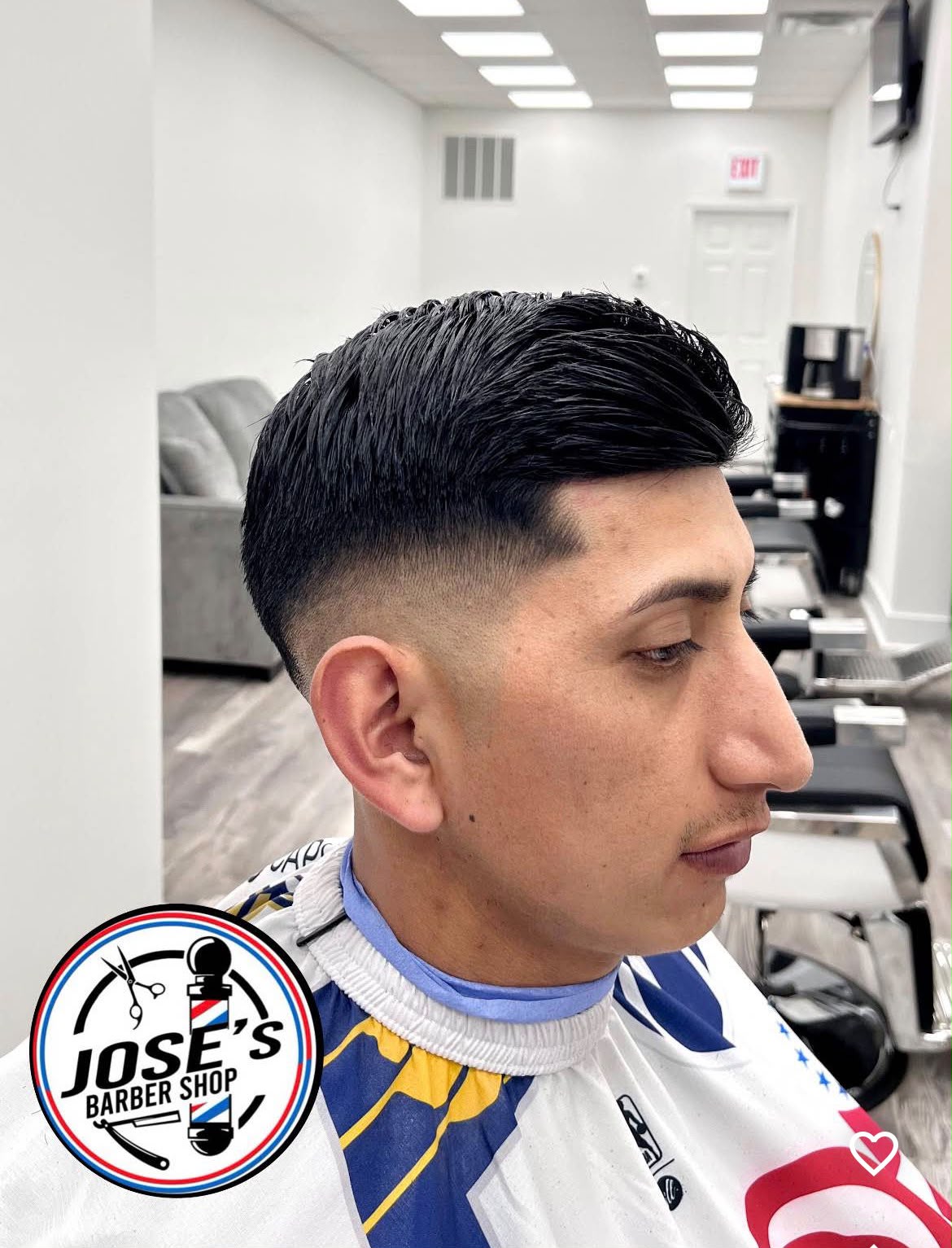 Un corte peinado de lado sobre un skin fade medio con un delineado marcado en el nacimiento del cabello, de perfil, con el sofá de espera y las sillas de barbero al fondo