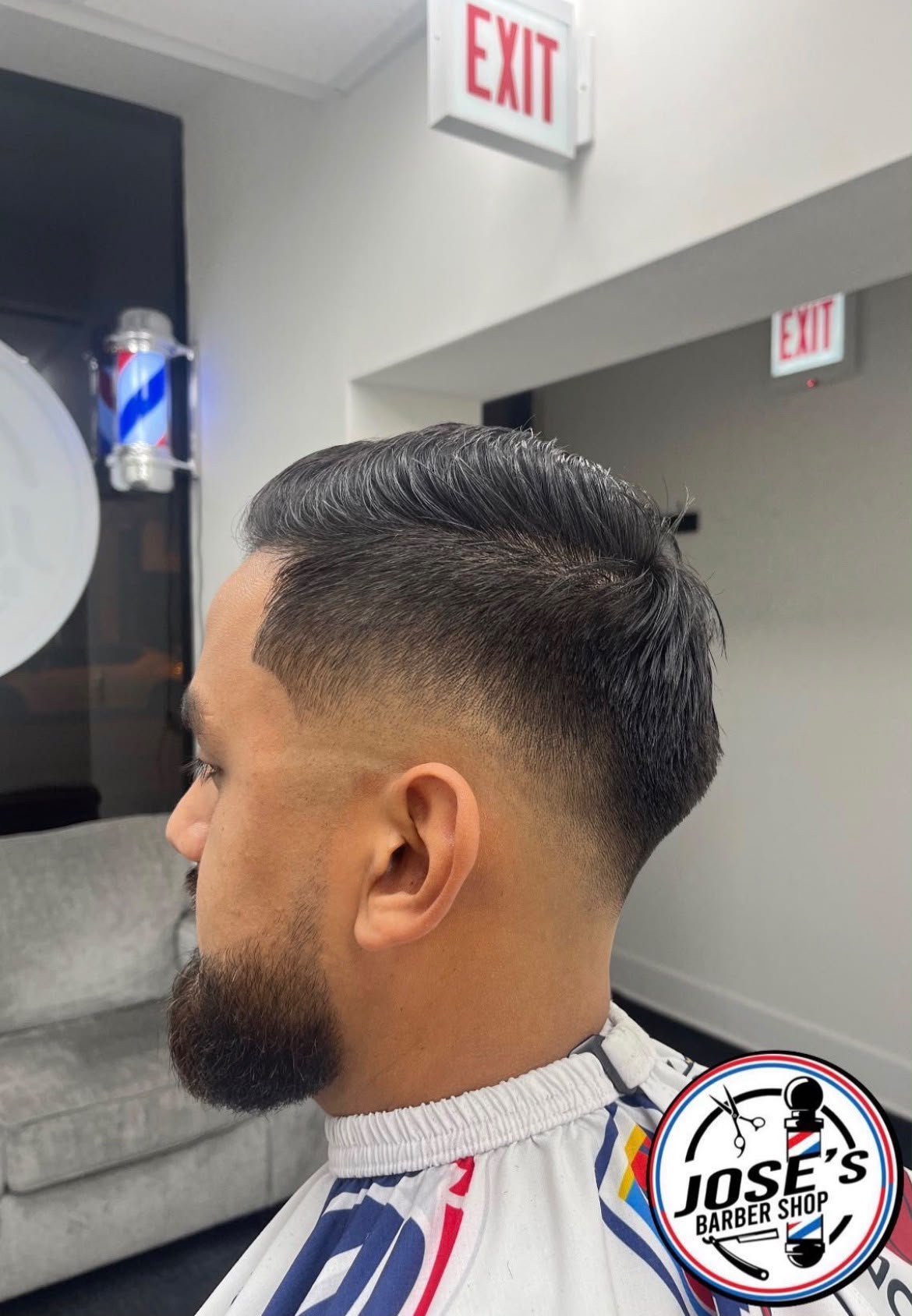 Un peinado de lado con raya marcada a navaja sobre un skin fade medio, rematado con una barba completa perfilada y delineada en la mandíbula, fotografiado frente al poste de barbero iluminado de la barbería