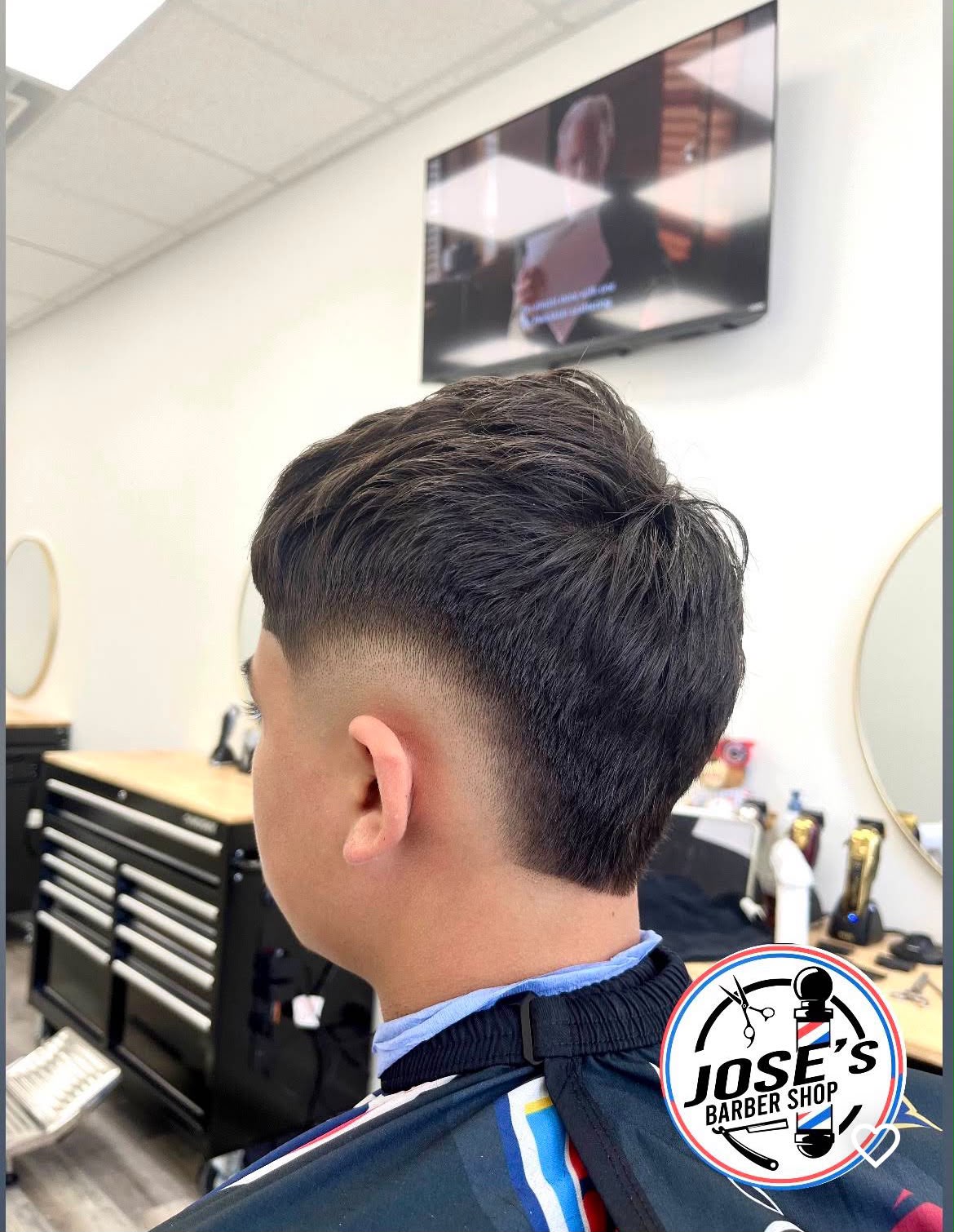 Cabello oscuro y abundante, con textura arriba y más largo en la parte de atrás, con un taper fade bajo detrás de la oreja, fotografiado desde atrás con la televisión de pared y los espejos redondos de la barbería al fondo