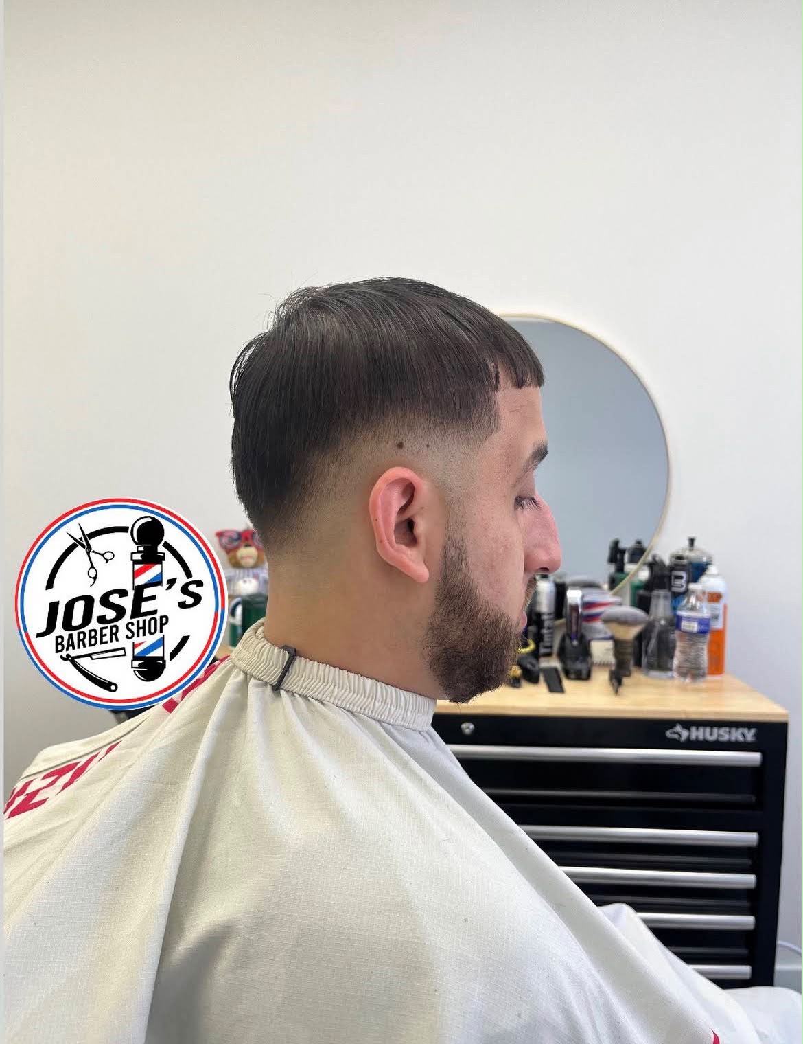 Un flequillo recto sobre un skin fade bajo en los lados y la nuca, rematado con una barba completa recortada y delineada, de perfil junto al espejo redondo y el gabinete de herramientas de la barbería