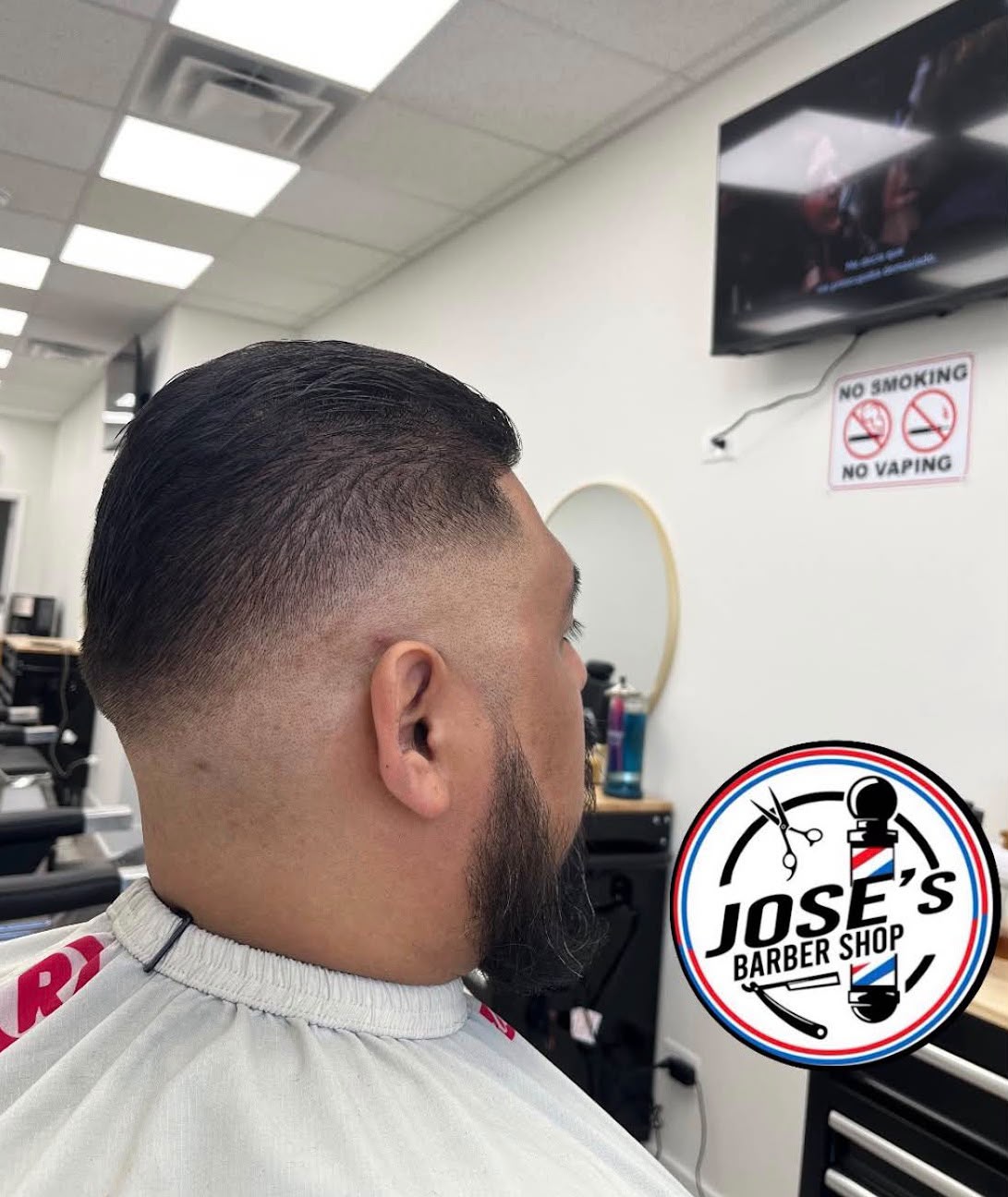 Cabello oscuro peinado hacia atrás y liso arriba sobre un taper fade bajo, con una patilla larga y la barba perfilada en la mandíbula, visto desde atrás con la televisión y un espejo redondo en la pared