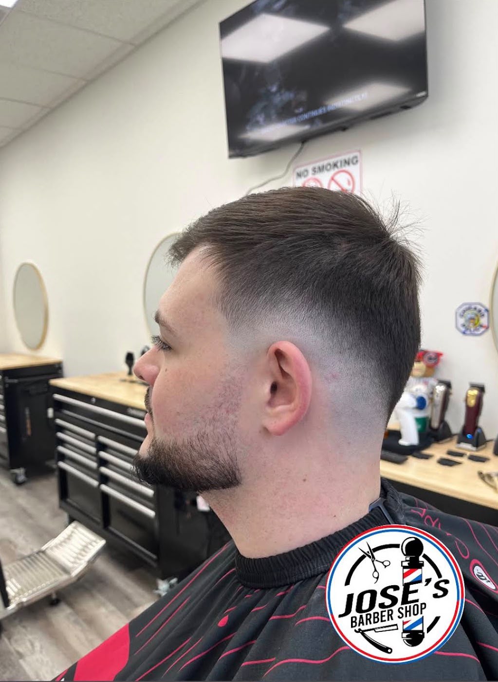 Un corte corto peinado hacia atrás sobre un skin fade medio con una línea limpia en la sien, junto con una perilla recortada, con las máquinas del barbero alineadas en la mesa al fondo