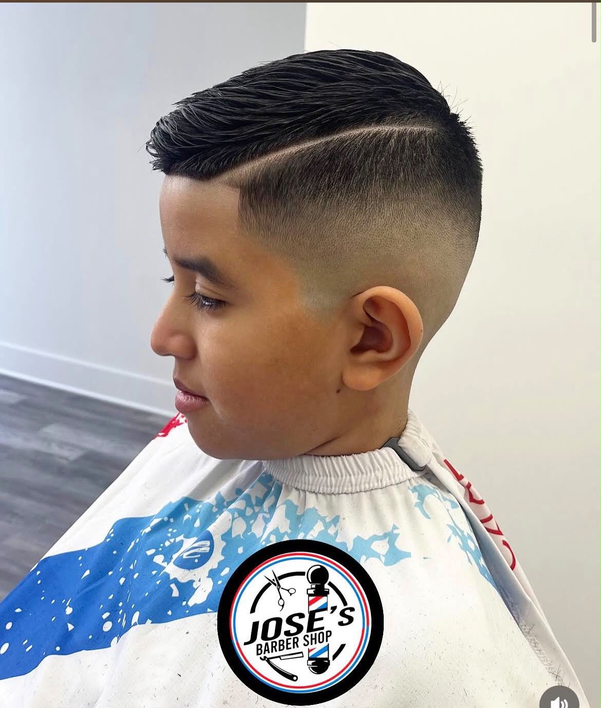 El corte de un niño peinado de lado con raya marcada a navaja y un skin fade alto en los lados, terminado en la silla bajo una capa azul y blanca