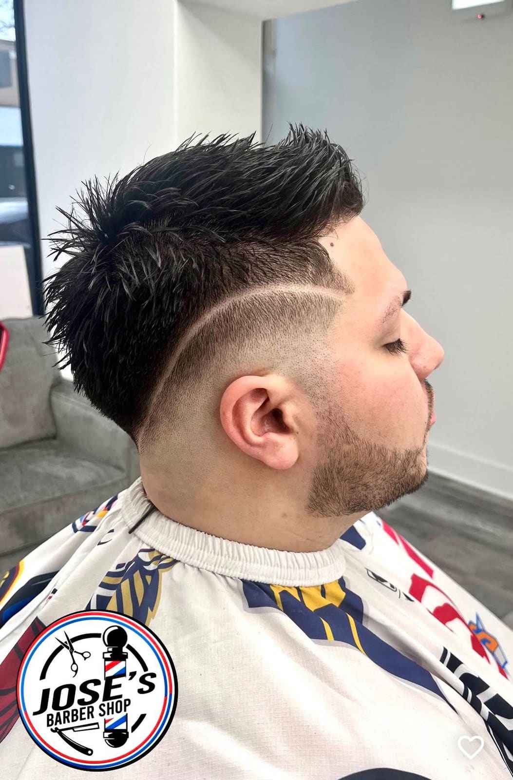 Cabello negro levantado y con puntas arriba, con una línea marcada a navaja y un skin fade alto en el lado y una barba corta recortada, fotografiado junto al sofá de espera de la barbería