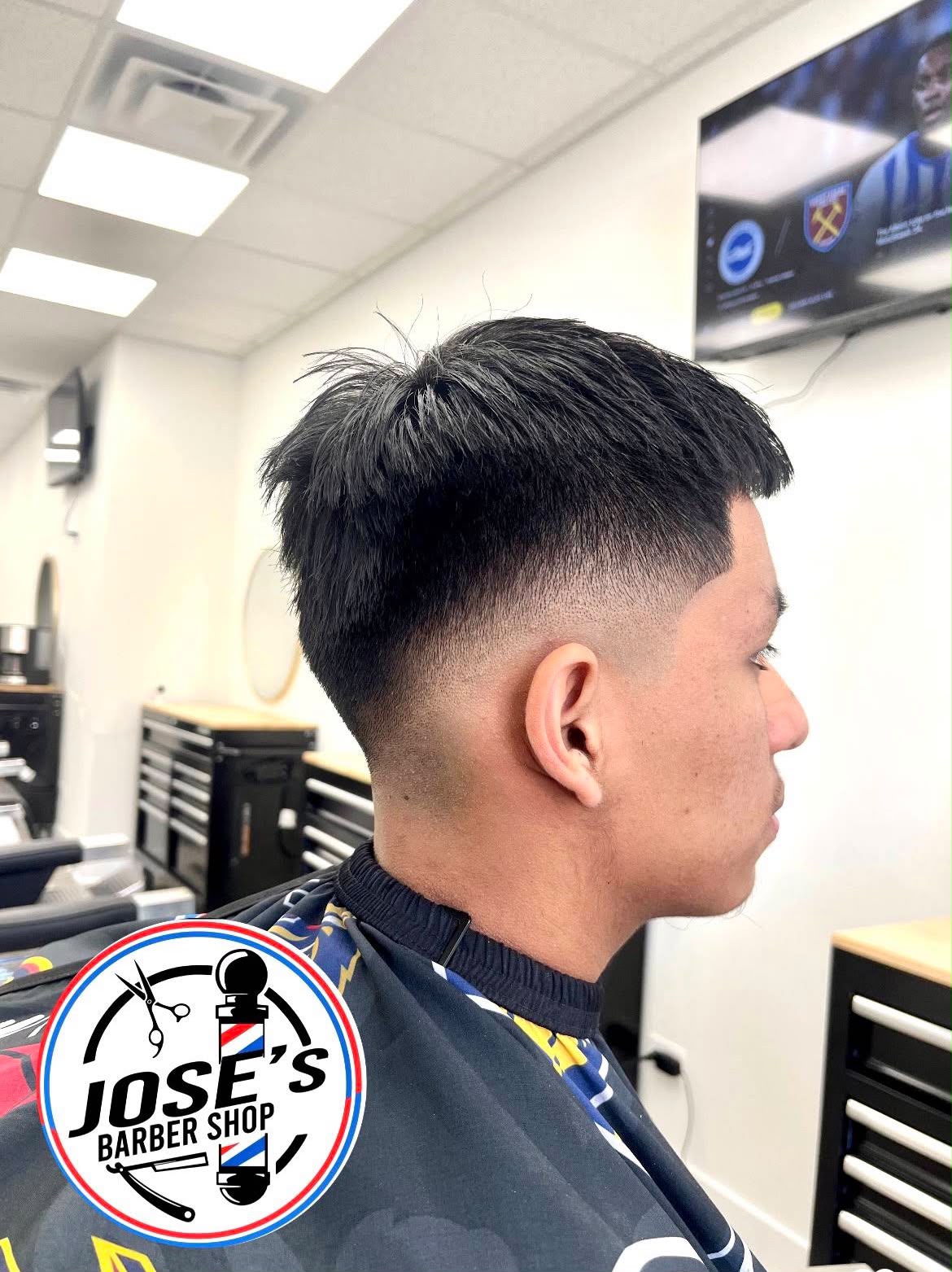 Cabello negro abundante, largo y con textura arriba, sobre un skin fade medio y un delineado marcado en la sien, de perfil con la televisión y las estaciones de barbero al fondo