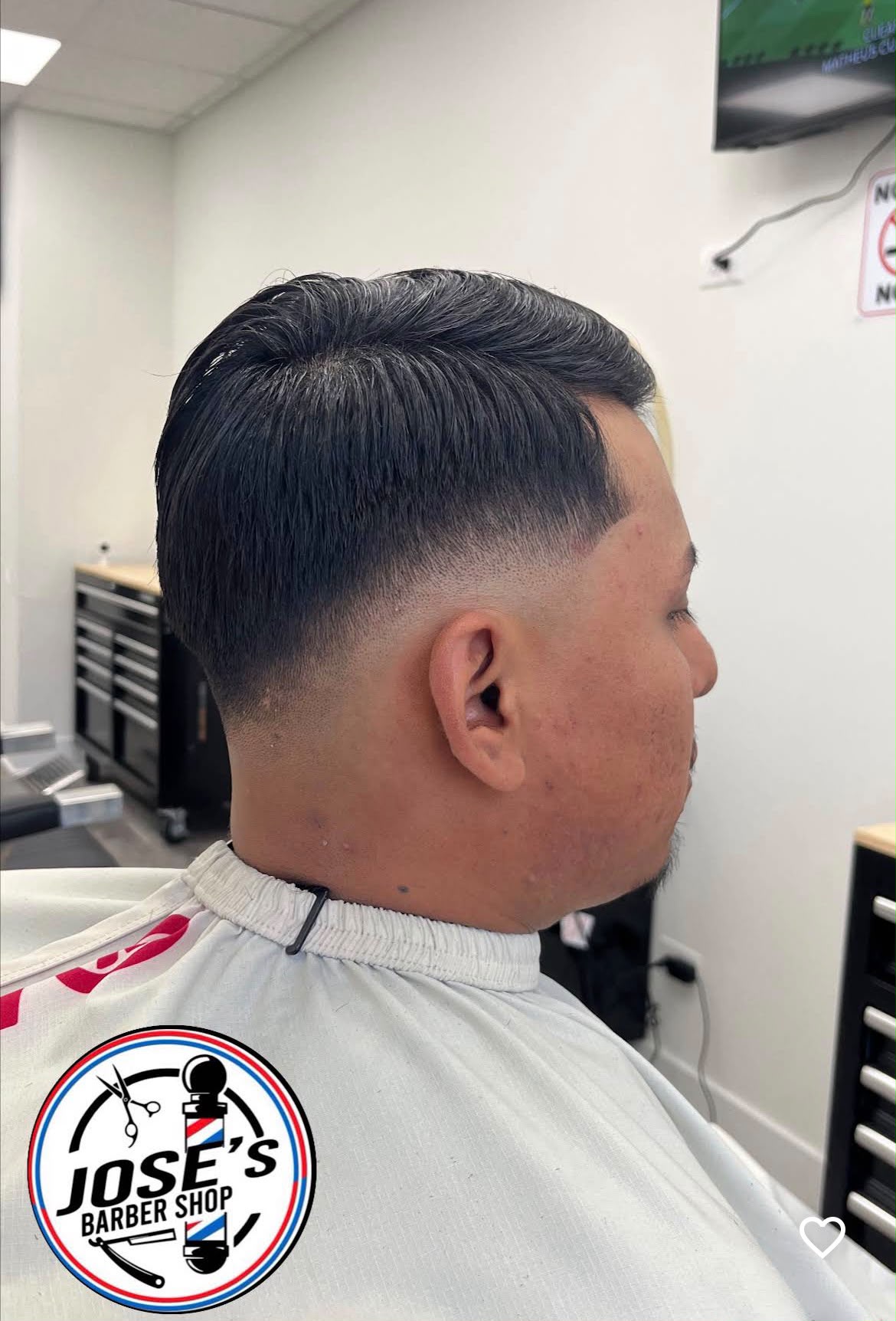 Cabello oscuro peinado hacia un lado y abundante arriba, difuminado en un taper fade bajo alrededor de la oreja y en el cuello, visto desde atrás en la silla