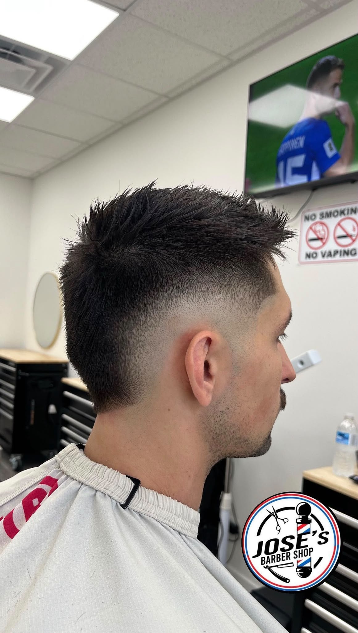 Un corte corto con textura, levantado arriba sobre un skin fade medio, difuminado hasta el cuello y con bigote y barba recortada, con la televisión de pared y un espejo redondo de la barbería al fondo